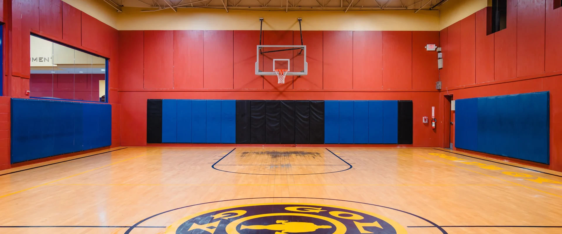 A basketball court with polished wood flooring, featuring a central logo of "Gold's Gym" in yellow and blue. Red and blue padded walls add vibrancy. The hoop is centered against the red wall, conveying an energetic and professional sports environment.