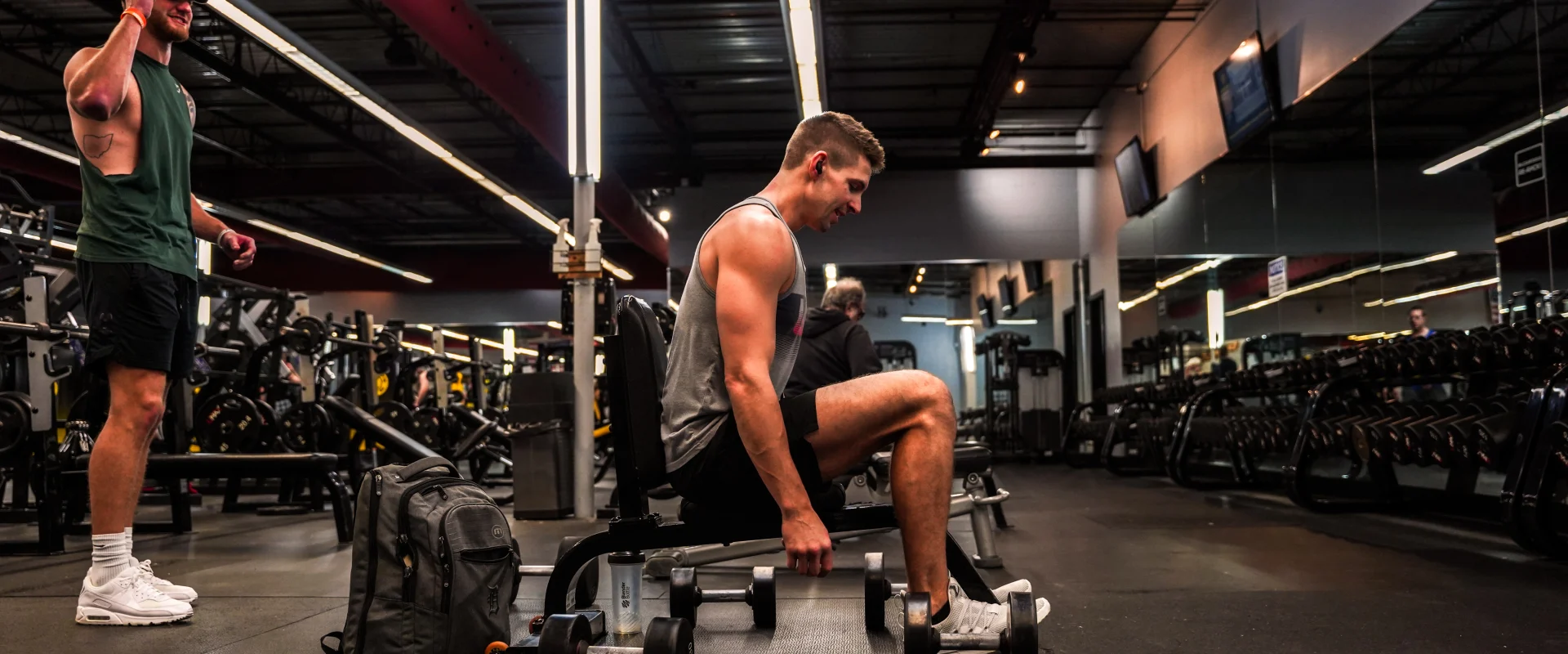 A man seated on a bench at Gold’s Gym Bellevue, Nashville, Tennessee, holding dumbbells in each hand during a free weight workout. Nearby, another man stands wearing a green tank top and a cap. The gym features rows of dumbbells, mirrored walls reflecting the equipment and members, and industrial-style lighting with red accents along the ceiling. A gray gym bag and water bottle rest on the floor in front of the seated man.