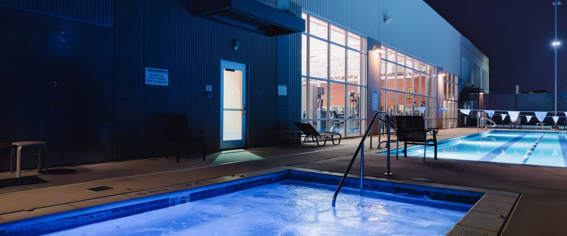 Nighttime shot of illuminated hot tub and pool beside a modern building. The blue-lit water contrasts with the dark sky, creating a serene ambiance.
