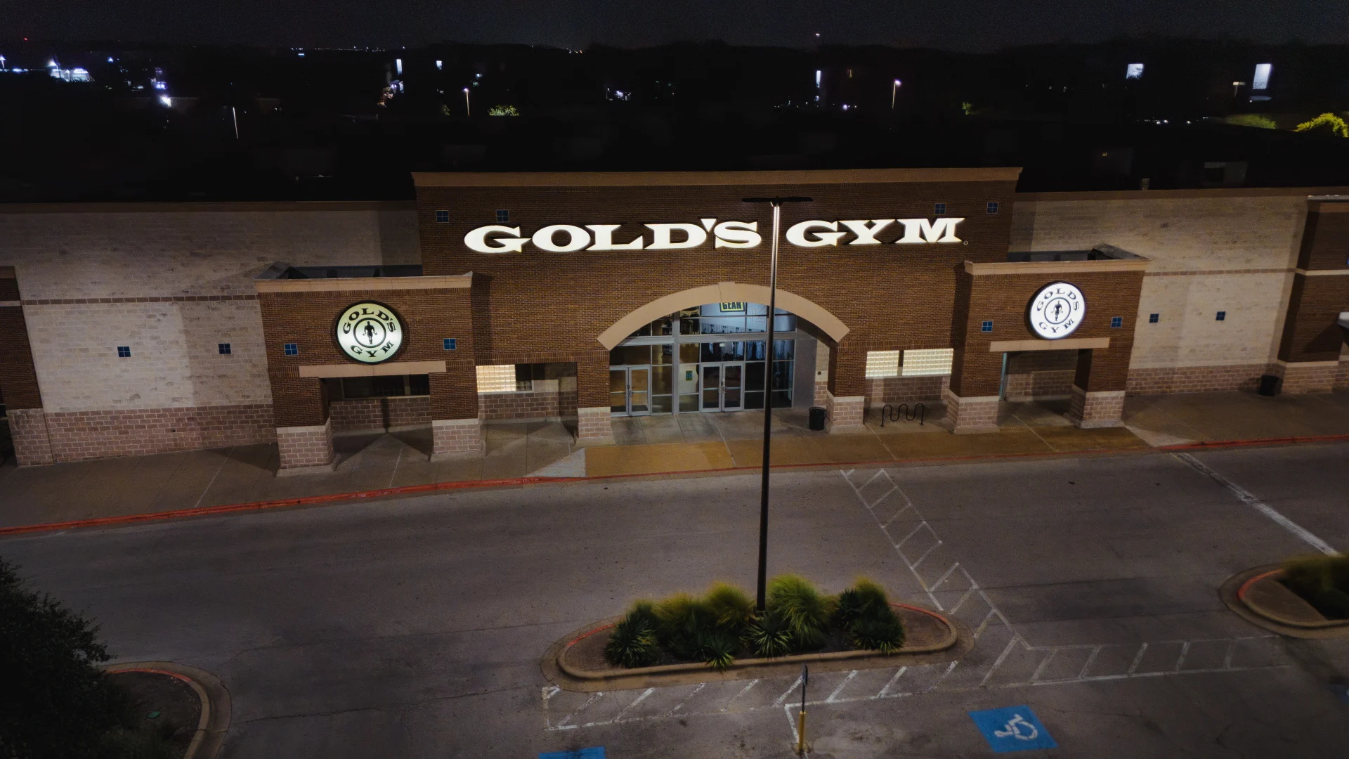 Nighttime view of Gold’s Gym Austin (Anderson Arbor) exterior featuring a large illuminated “Gold’s Gym” sign above the main glass entrance doors. The brick facade includes circular Gold’s Gym logos on the walls to the left and right of the entrance.