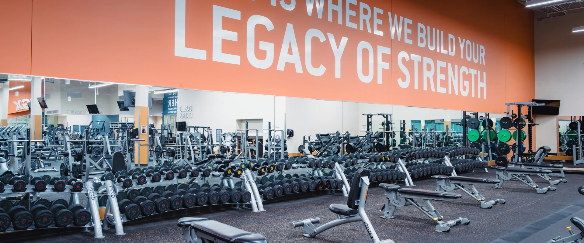 A modern gym with an orange wall featuring the bold text, "This is where we build your legacy of strength." Rows of weights and benches fill the well-lit space.