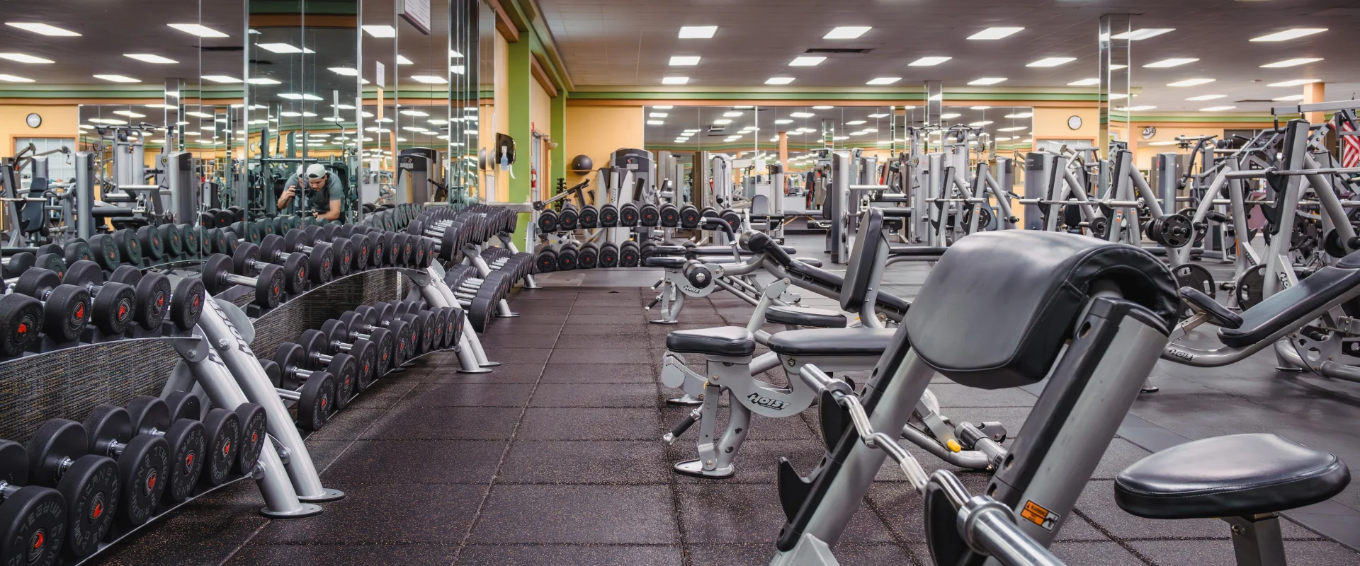 Empty gym with rows of dumbbells, weight benches, and exercise machines. Mirrors line the walls, creating a spacious and organized atmosphere.