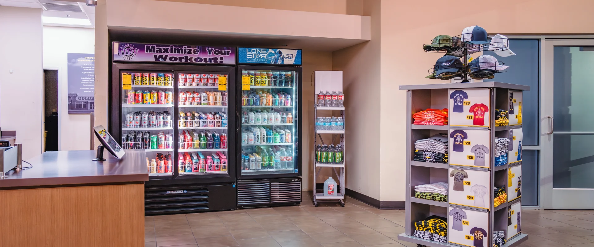 Pro shop in Austin (Westlake), featuring a beverage cooler stocked with workout drinks and supplements, alongside a display rack with Gold's Gym branded apparel, including hats, t-shirts, and gym gear. The tiled floor and clean layout create an organized and welcoming retail space.