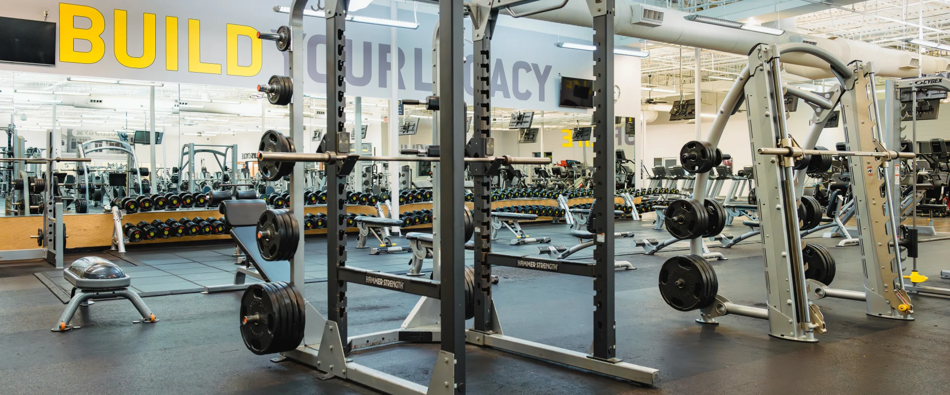 Free weight area at Gold's Gym in Austin (Hesters Crossing), featuring squat racks, a smith machine, and a variety of weightlifting equipment neatly arranged on black rubber flooring. The space is brightly lit with high ceilings, mirrored walls reflecting rows of dumbbells and benches, and motivational text reading "BUILD YOUR LEGACY" prominently displayed on the wall.