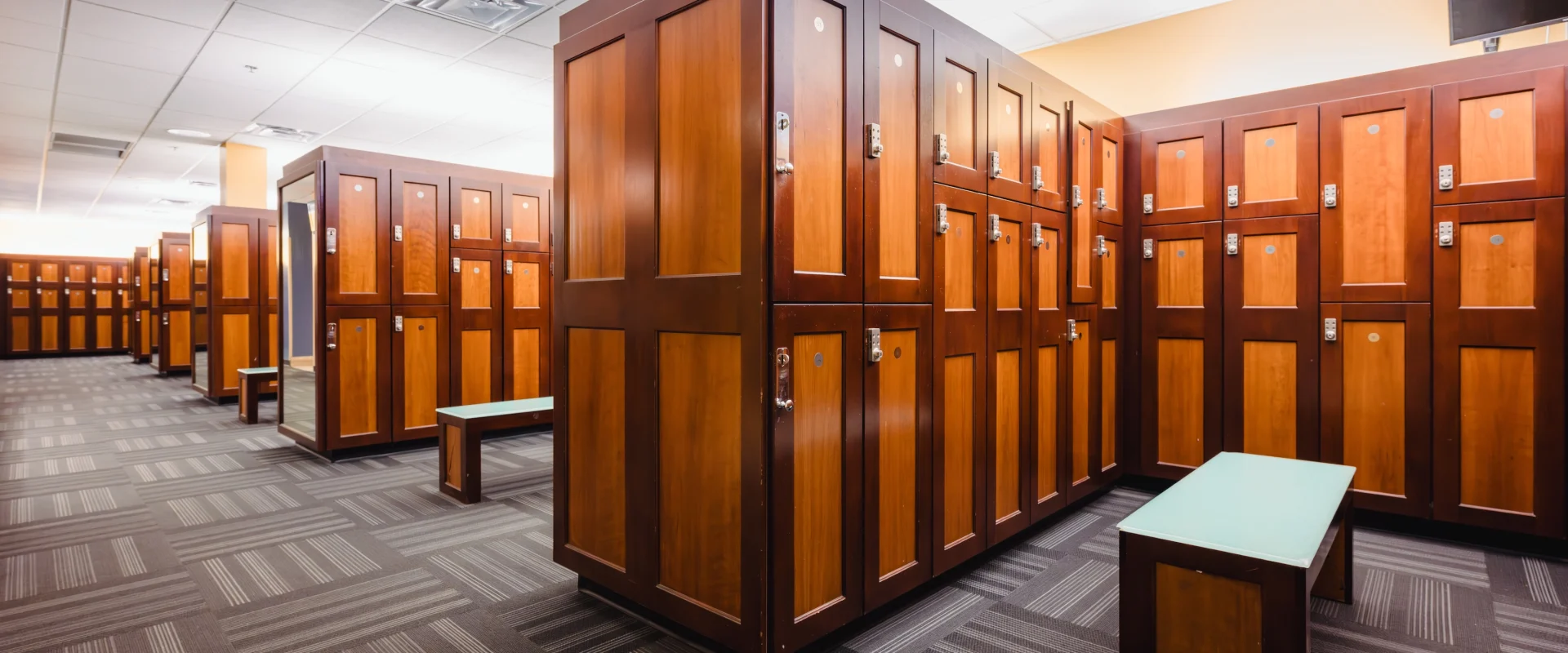 Modern locker room with rows of wooden lockers and blue benches on a carpeted floor. The setting is clean and organized, conveying a sense of order.