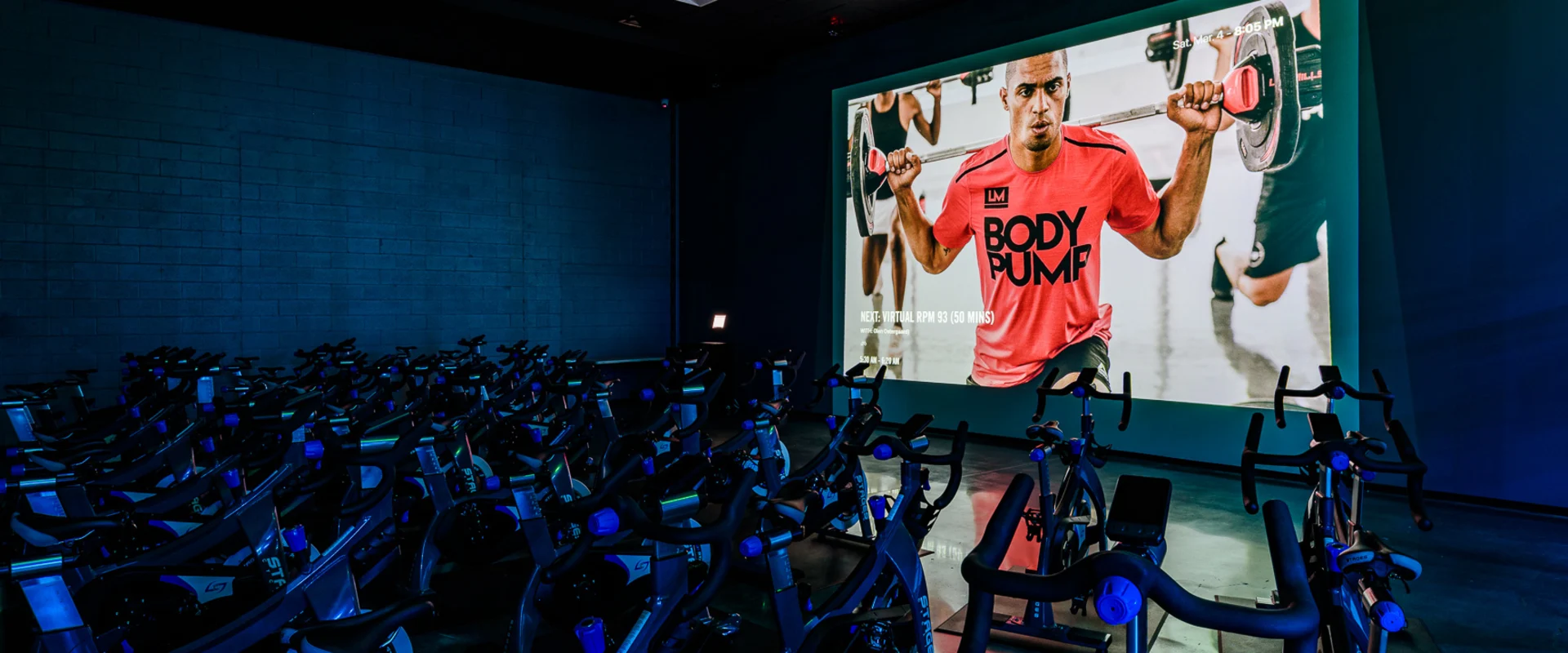 The group cycle studio at Gold’s Gym Cerritos features rows of stationary bikes arranged on a polished concrete floor. A large projector screen at the front displays a virtual workout with an instructor in a red 'Body Pump' shirt. Blue accent lighting casts a glow over the dark walls and ceiling, creating an immersive training environment.