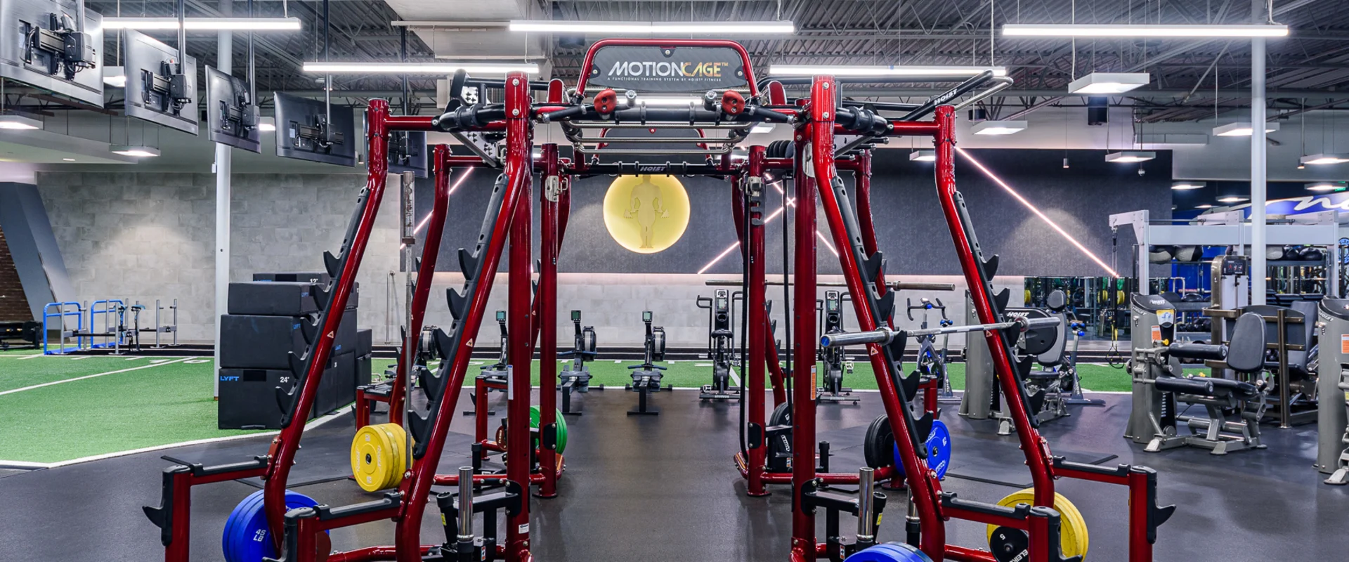 The power rack area features a bold red Motion Cage training system equipped with multiple squat stations, pull-up bars, and storage for color-coded weight plates. The black rubber flooring contrasts with the turf section in the background. Overhead LED lighting illuminates the space, while mounted TVs line the ceiling. A Gold’s Gym logo glows on the back wall, reflecting off the sleek black and gray walls.