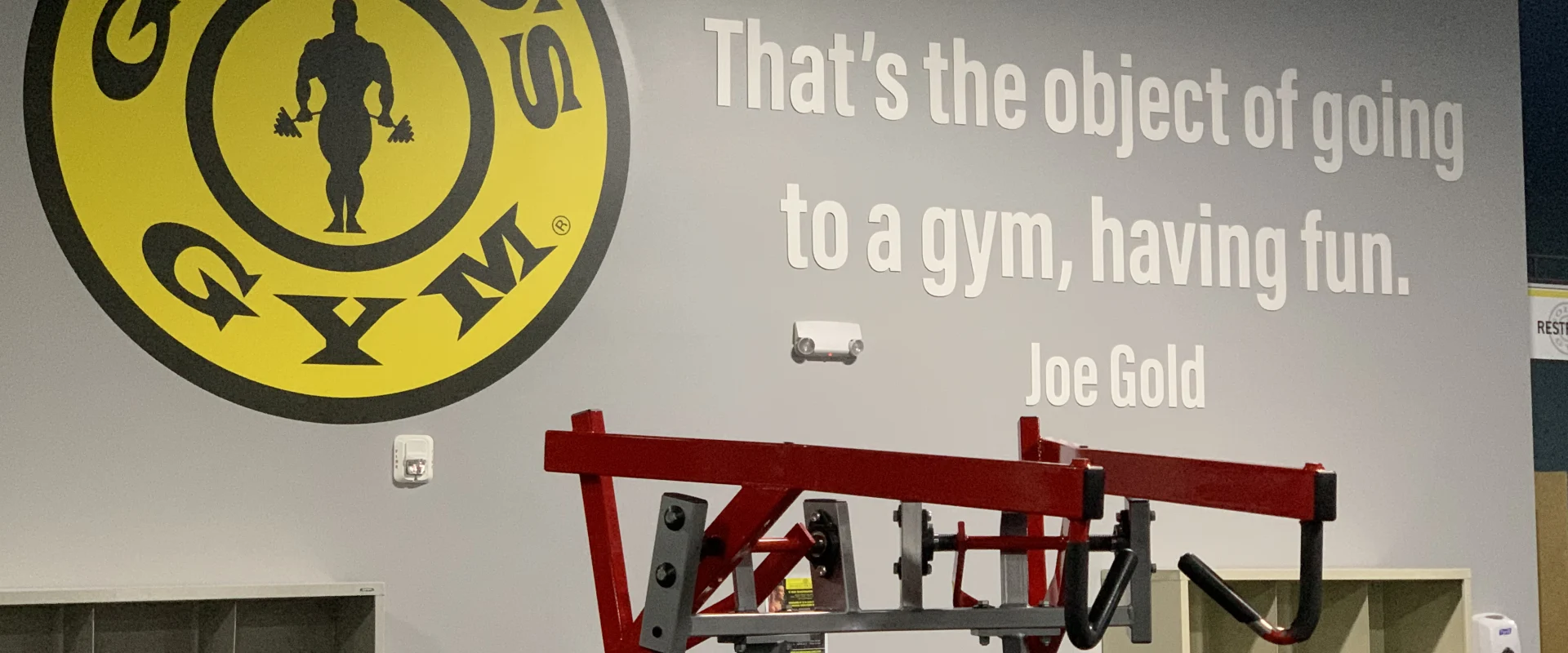 A large yellow circular logo with the text "GOLD'S GYM" is prominently displayed on the wall, alongside a quote attributed to Joe Gold stating "That's the object of going to a gym, having fun." In the foreground, various gym equipment and storage shelves are visible.