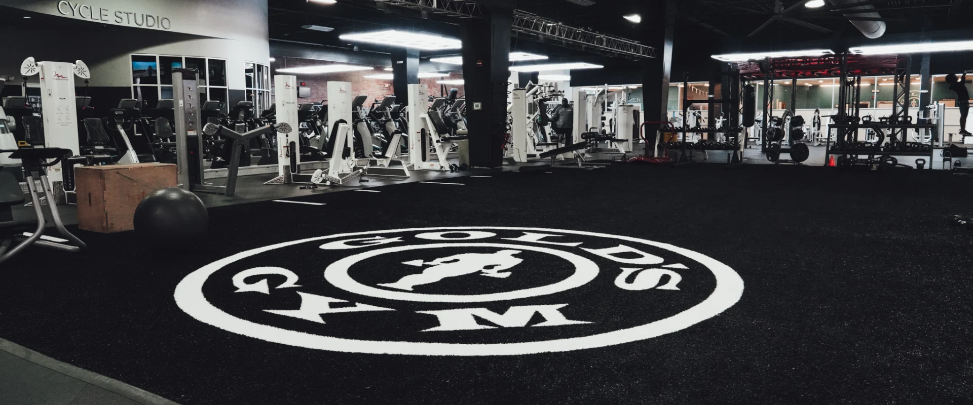 A functional training zone at Gold's Gym Brassfield, featuring a black turf area with the Gold’s Gym logo in the center. The space is surrounded by cable machines, squat racks, and cardio equipment. A wooden plyometric box and medicine ball rest on the turf, ready for performance training.