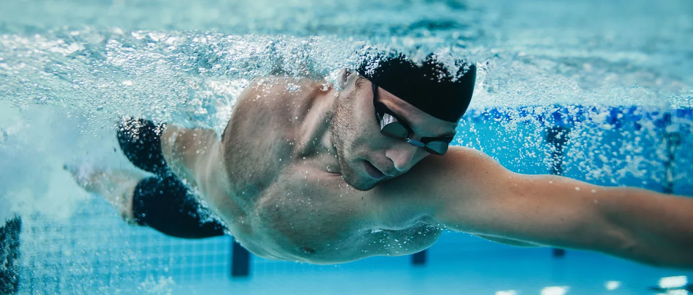 A person wearing swimming goggles is submerged underwater, with water splashing around them against a blurred blue background.
