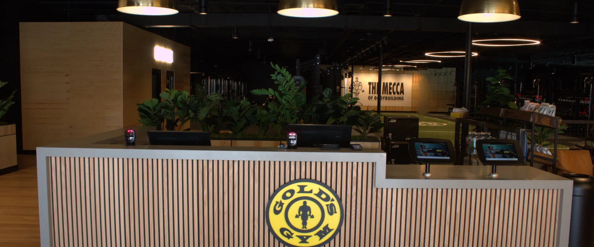 Front desk at Gold's Gym Austin, Burnet Rd featuring vertical wood slats, gold pendant lights, lush greenery, and a view of the gym floor and turf area in the background.