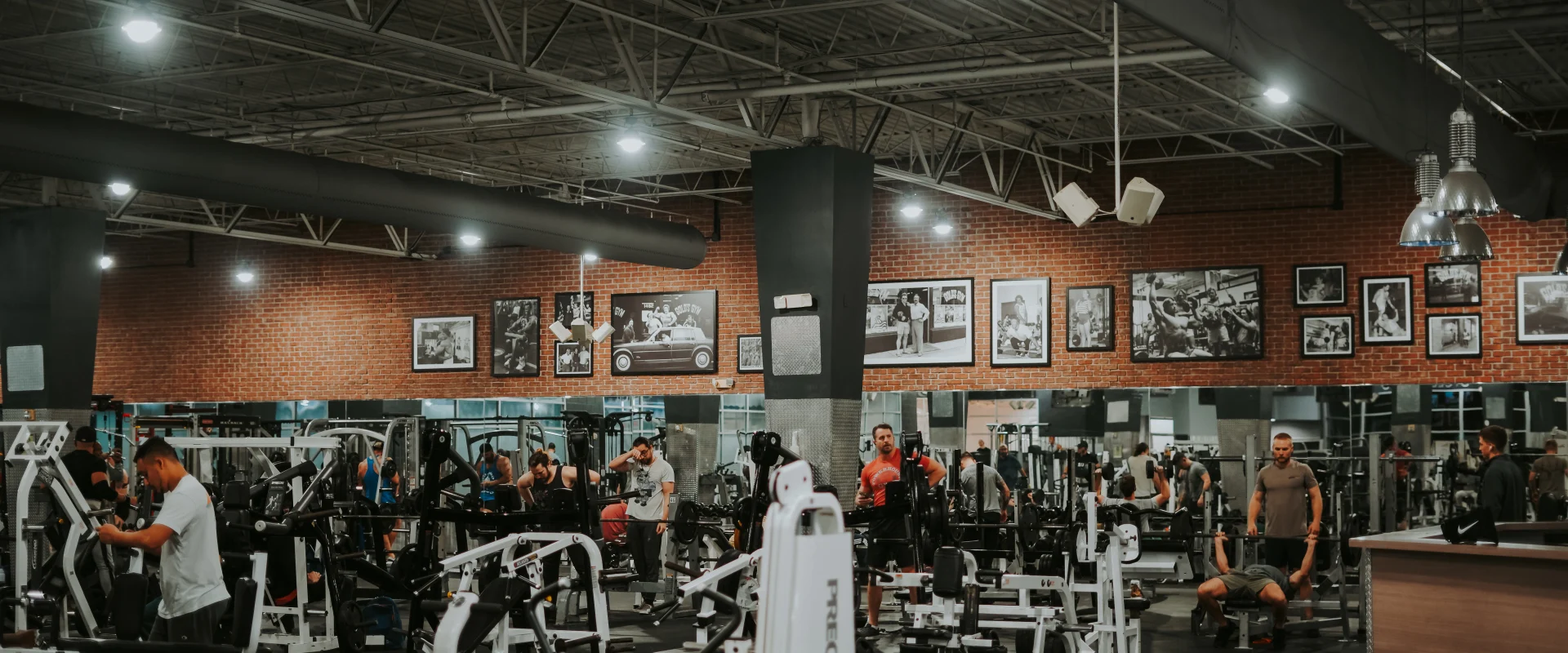 Machine weights area at Gold’s Gym Tennessee, Lee Highway, featuring rows of resistance machines and equipment arranged under a high ceiling with exposed beams and ducts. The brick accent wall in the background is adorned with framed historical photos, adding a classic touch to the space. Overhead lighting illuminates the area, while gym members actively engage in their workouts.