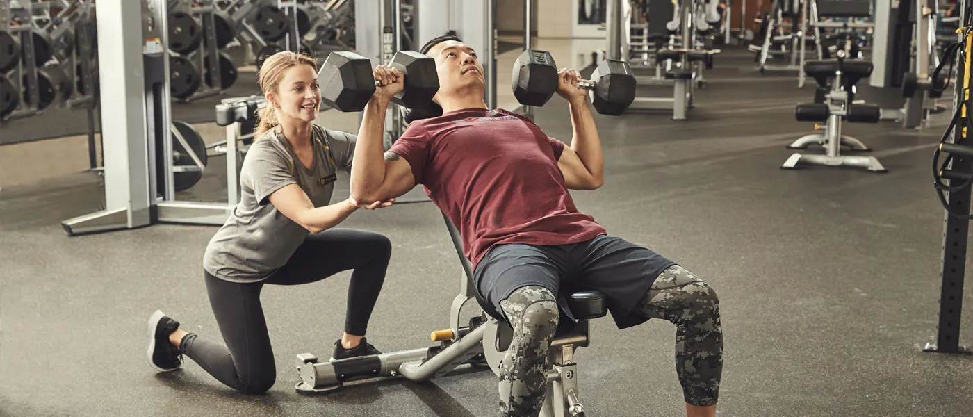 Two people, a man and a woman, are working out together in a gym setting. The man is using a weight machine while the woman is assisting him.