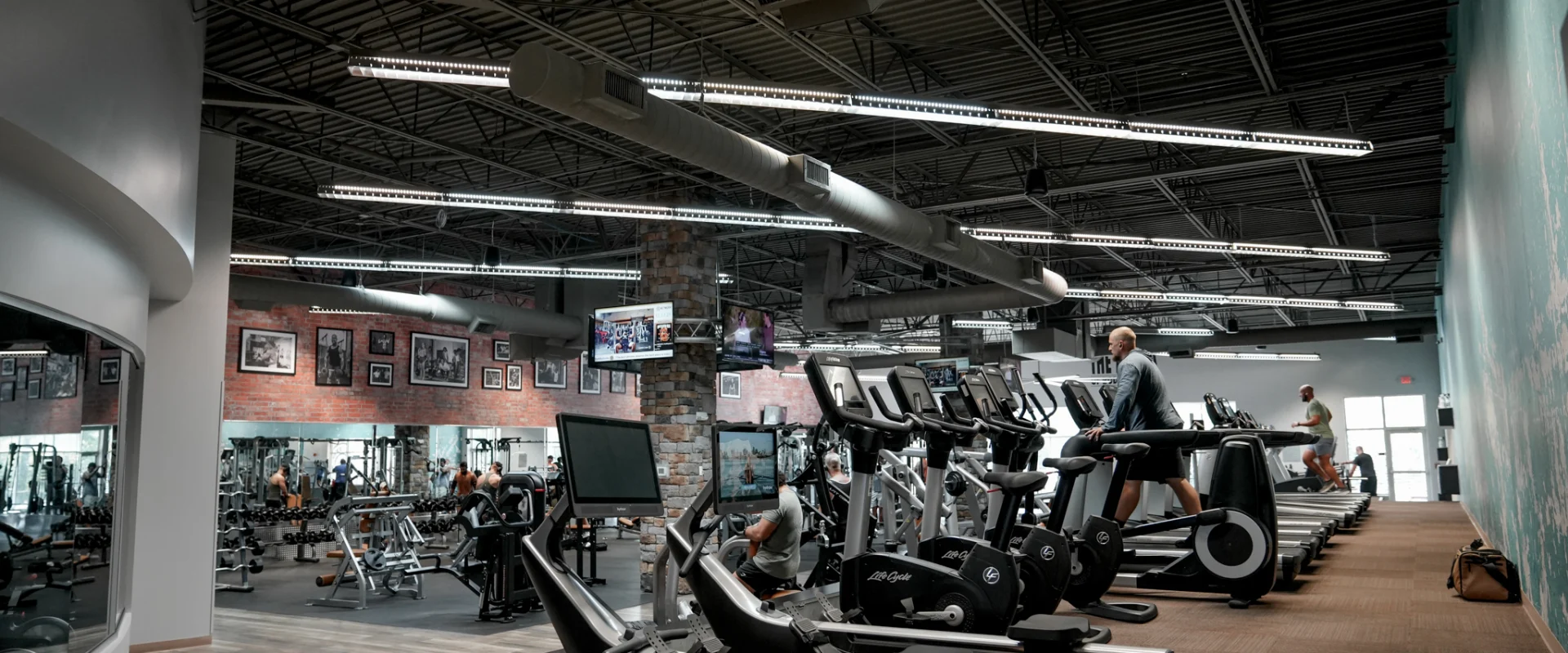 A spacious gym floor at Gold's Gym Rock Hill, showcasing a variety of cardio and strength equipment. Treadmills, ellipticals, and weight machines line the space, with stone columns and industrial lighting enhancing the aesthetic. Large mirrors and brick walls contribute to the gym’s classic bodybuilding atmosphere.