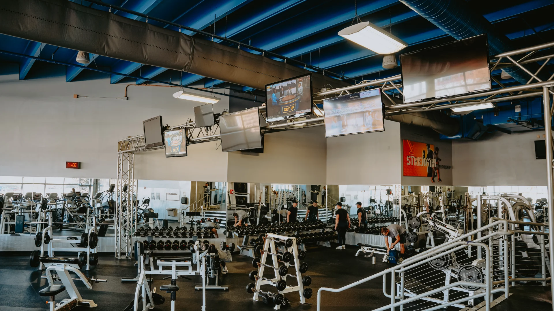 Dumbbell area at Gold’s Gym Tennessee, Downtown Chattanooga, featuring a neatly arranged row of dumbbells on racks against mirrored walls, creating an expansive feel. The space includes a dark rubber floor, high ceiling with exposed blue ducts, and overhead lighting. Mounted televisions and a motivational “Strength” sign enhance the workout environment, with reflections of the gym space visible in the mirrors.