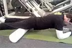 A person in black clothing is lying on a green exercise mat in what appears to be a gym or fitness facility, with various exercise equipment visible in the background.