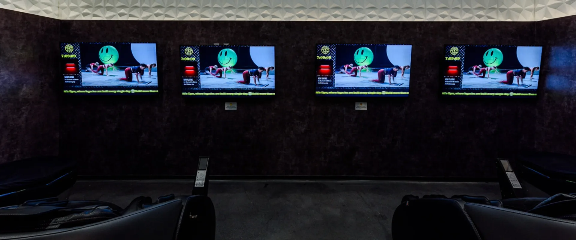 Recovery area at Gold’s Gym Long Beach, designed for post-workout relaxation. Black massage chairs are positioned in front of a dark-textured wall, each facing a mounted screen displaying guided recovery and wellness content. The room features a sleek white geometric ceiling design, soft ambient lighting, and a calming atmosphere for members to unwind and recharge.