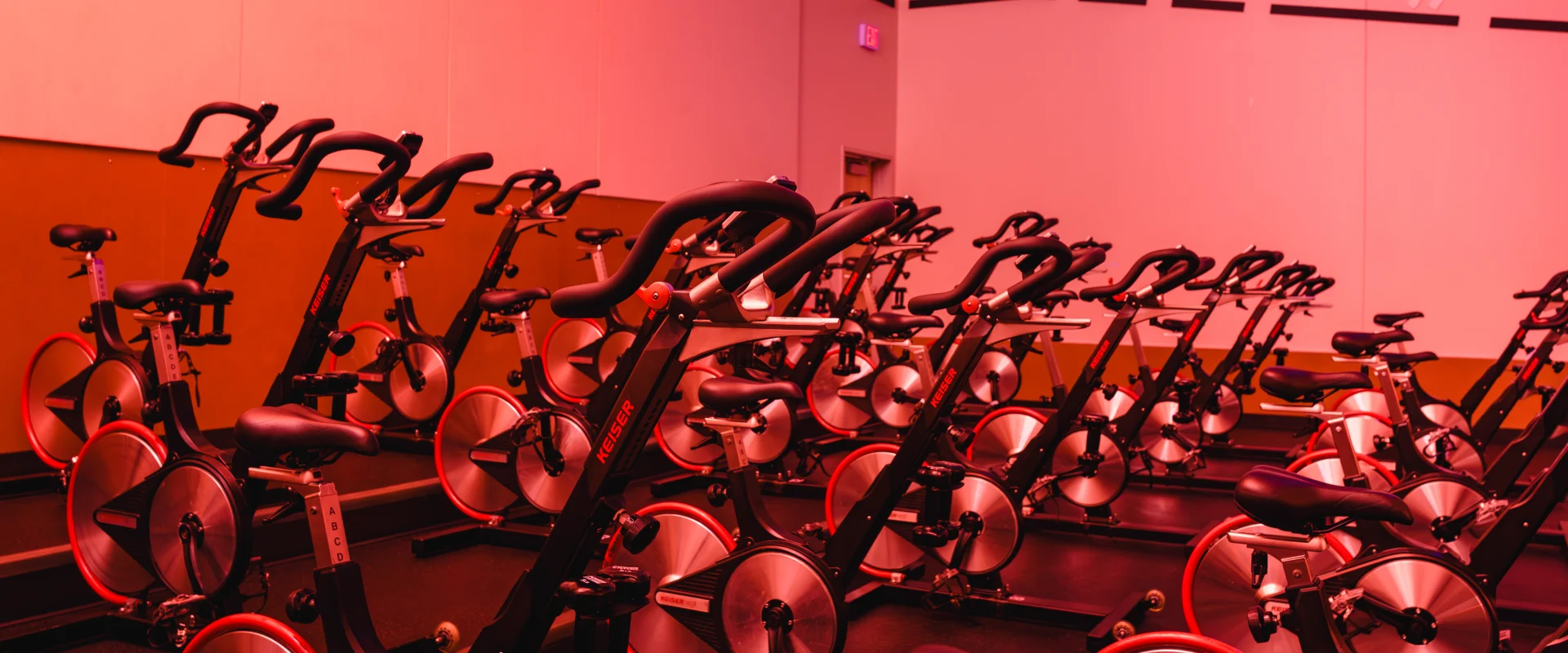 A cycling studio illuminated by red lighting, featuring rows of Keiser stationary bikes arranged on tiered platforms in a sleek, modern setting.