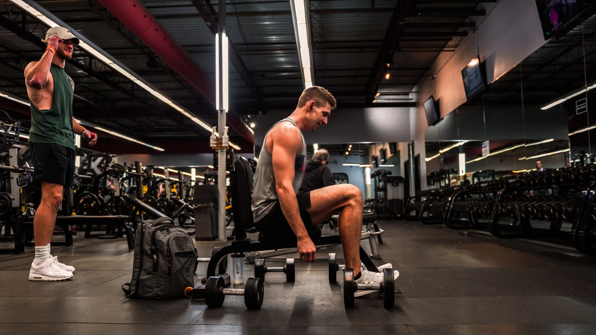 A man seated on a bench at Gold’s Gym Bellevue, Nashville, Tennessee, holding dumbbells in each hand during a free weight workout. Nearby, another man stands wearing a green tank top and a cap. The gym features rows of dumbbells, mirrored walls reflecting the equipment and members, and industrial-style lighting with red accents along the ceiling. A gray gym bag and water bottle rest on the floor in front of the seated man.