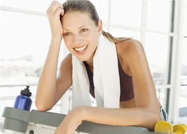 A smiling woman in workout clothes stands in front of a window, holding a towel and water bottle.