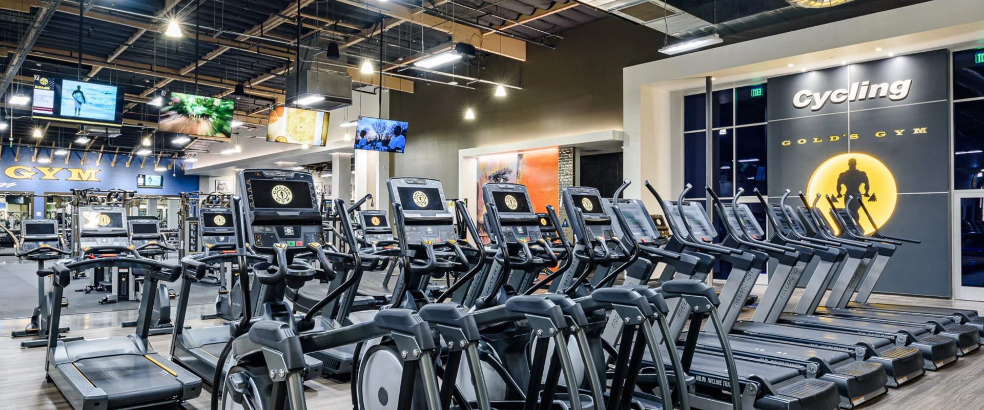 The cardio area at Gold’s Gym Cerritos features rows of treadmills and elliptical machines with digital screens, arranged on wood-style flooring. Large TVs hang from the ceiling, displaying various channels. The background includes a "Cycling" studio sign with the Gold’s Gym logo, along with exposed ductwork and industrial lighting overhead. The gym’s blue and yellow branding is visible on the back wall.