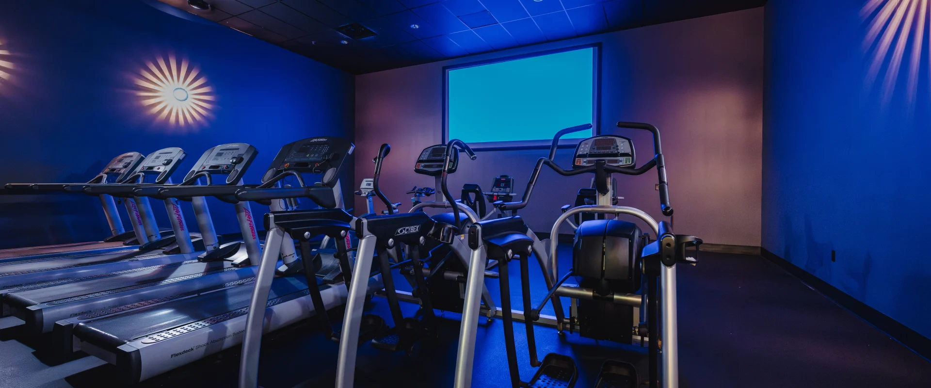 Cardio Cinema room at Gold’s Gym Austin (Anderson Arbor) featuring treadmills and elliptical machines facing a large projection screen. The room is dimly lit with wall-mounted lights creating a cinematic ambiance.