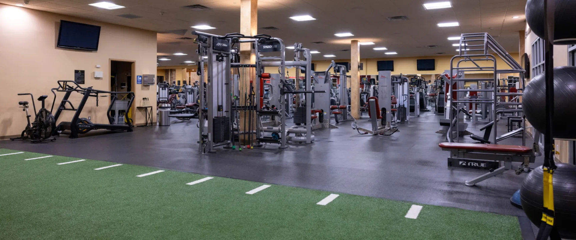 Turf training area at Gold’s Gym in East Northport, New York, featuring green artificial turf with white agility markings. Adjacent to the turf are functional training equipment, including battle ropes, medicine balls, and a TRX suspension system. The space opens into a larger gym area with strength machines, cardio equipment, and sled push tracks under bright overhead lighting.
