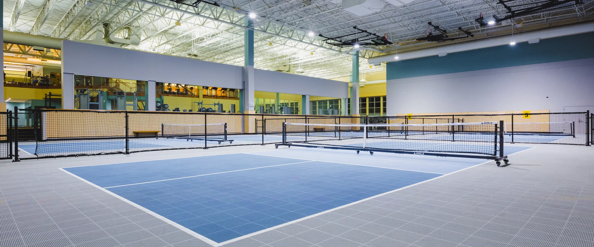 Indoor pickleball courts at Gold's Gym San Antonio Rogers Ranch, featuring two courts with a blue playing surface and a grey out-of-bounds area. The courts are separated by a net, with a glass window on one side overlooking the workout area. The space is enhanced by very high white ceilings and bright lighting, creating an open and well-lit environment