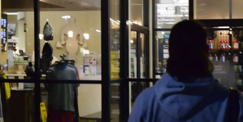 A person in a blue jacket stands in the foreground, with various items visible through the glass windows in the background.