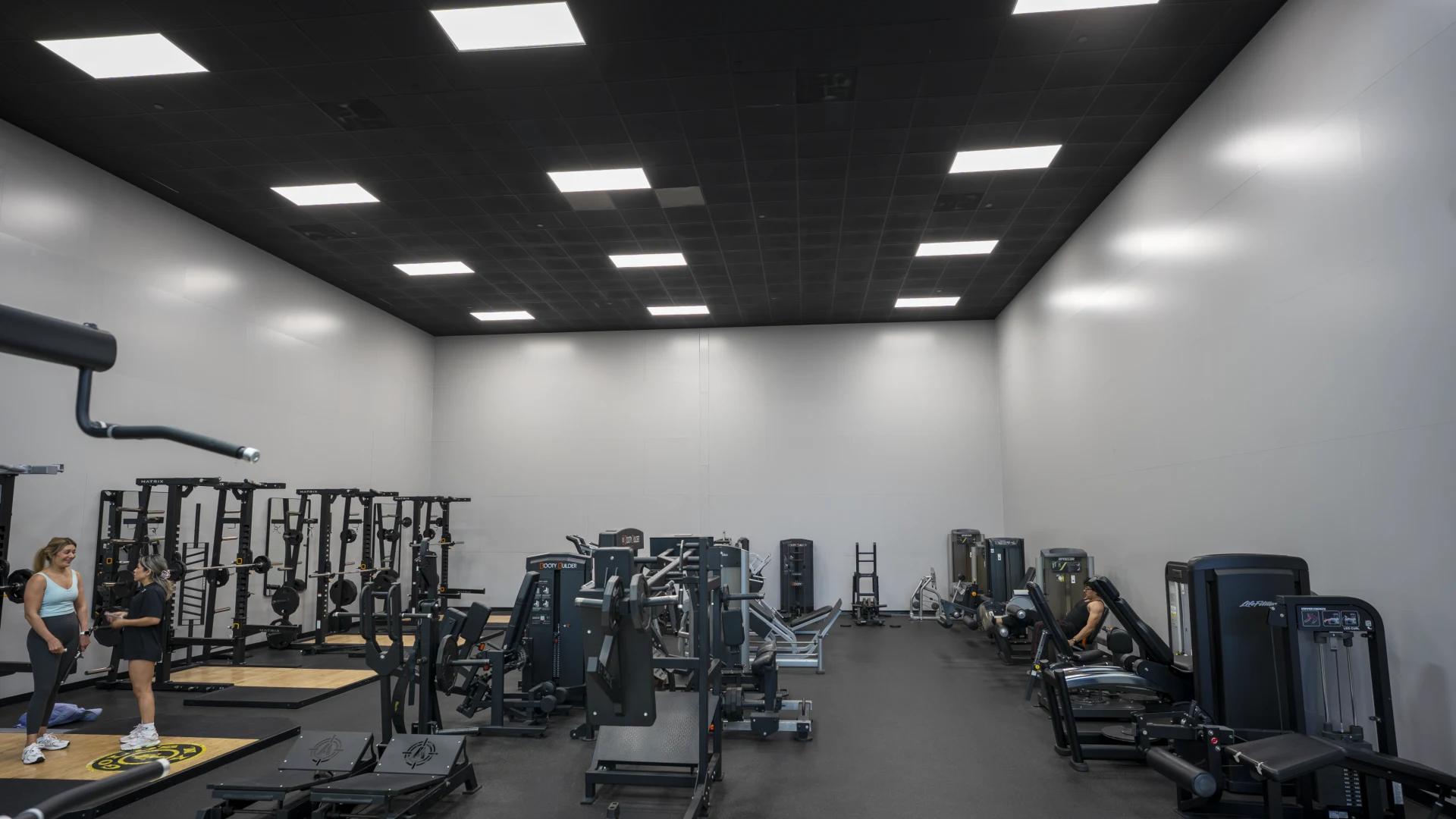 An expansive weight room floor featuring high ceilings with black tiles and rectangular LED panel lighting. The room is equipped with several squat racks, weightlifting platforms with Gold's Gym logos, and various selectorized strength machines. Two women are standing in the foreground on a lifting platform, and several other pieces of black gym equipment are distributed throughout the dark-floored space.