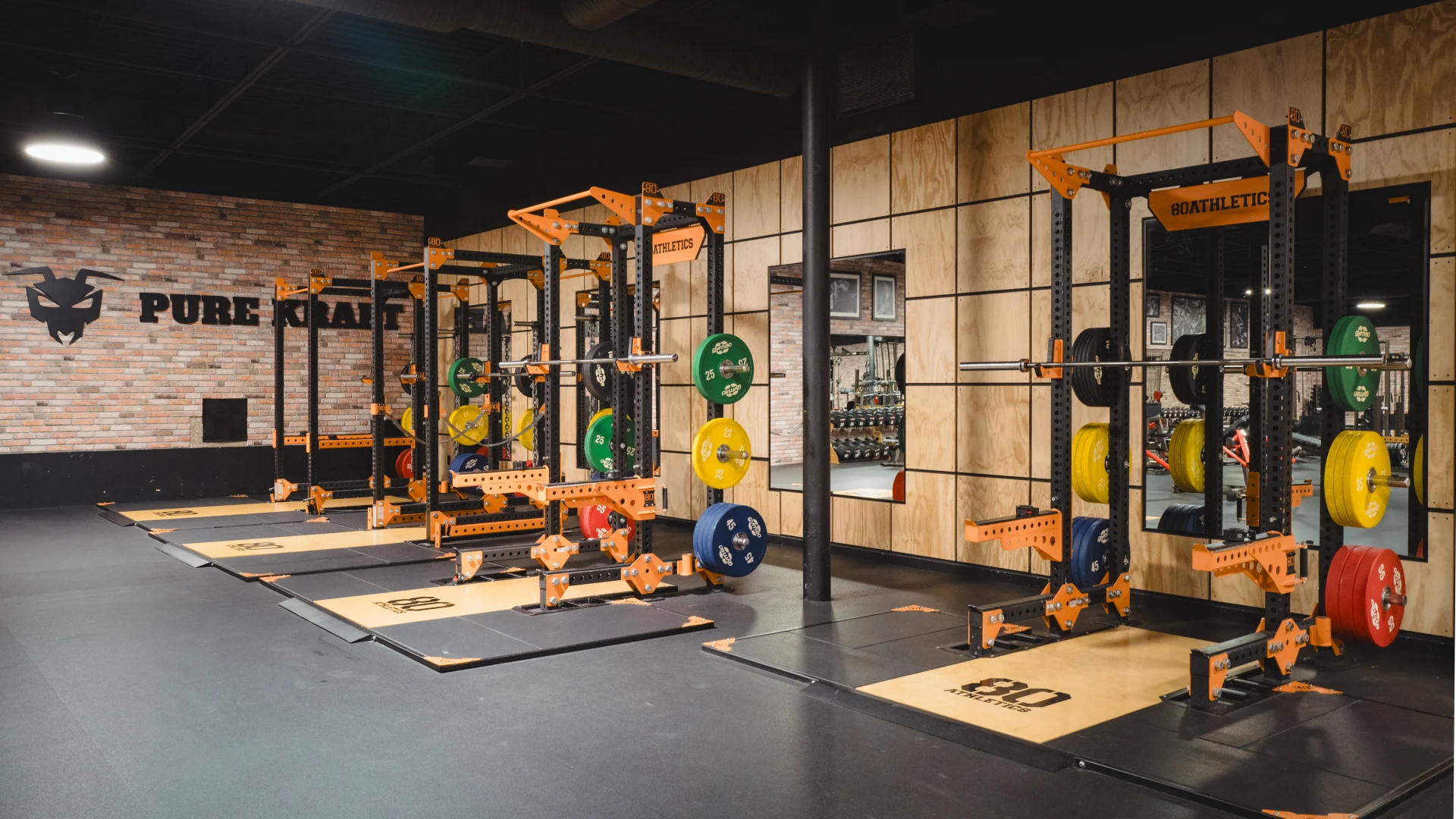 Power racks at Gold’s Gym Highland Meadows featuring orange frames and weight plates in multiple colors. The equipment sits on a rubber floor under ceiling lights, with a brick wall and the "Pure Kraft" logo in the background.