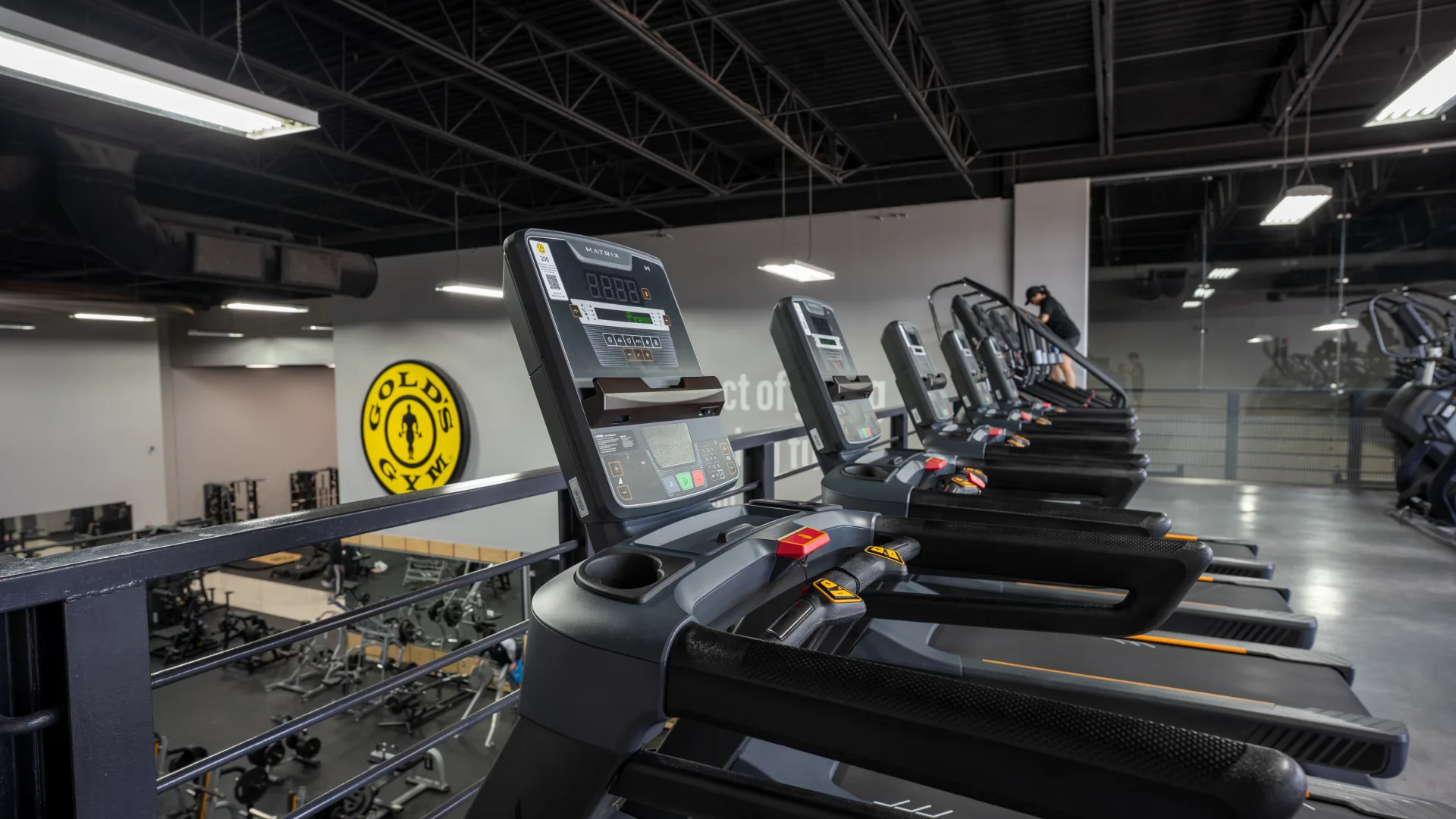 A row of Matrix brand treadmills lined up along a black metal mezzanine railing overlooking the main floor of a Gold’s Gym. The treadmills face away from the camera toward a grey wall featuring the circular yellow Gold’s Gym logo. Below the mezzanine, various weightlifting machines and equipment are visible on the lower level.