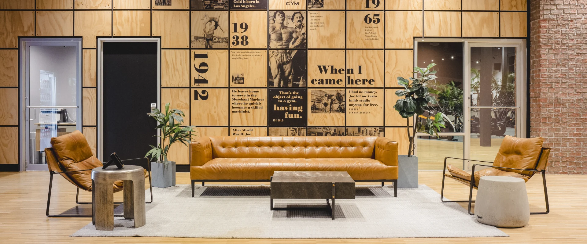 Modern lounge area at Gold’s Gym Little Elm featuring a caramel leather sofa, matching chairs, and a wooden accent wall displaying historical Gold’s Gym milestones and photos. A light rug, coffee table, and indoor plants enhance the warm, inviting atmosphere, with glass doors behind the lounge showcasing office space.