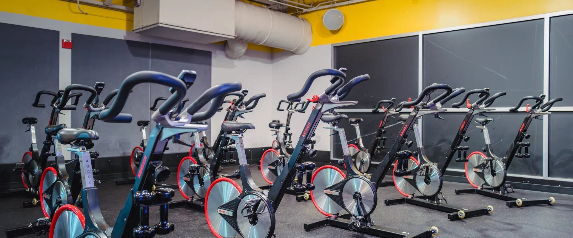 Cycle studio at Gold's Gym Live Oak, featuring rows of stationary bikes, shaded out windows, and bright yellow accents.