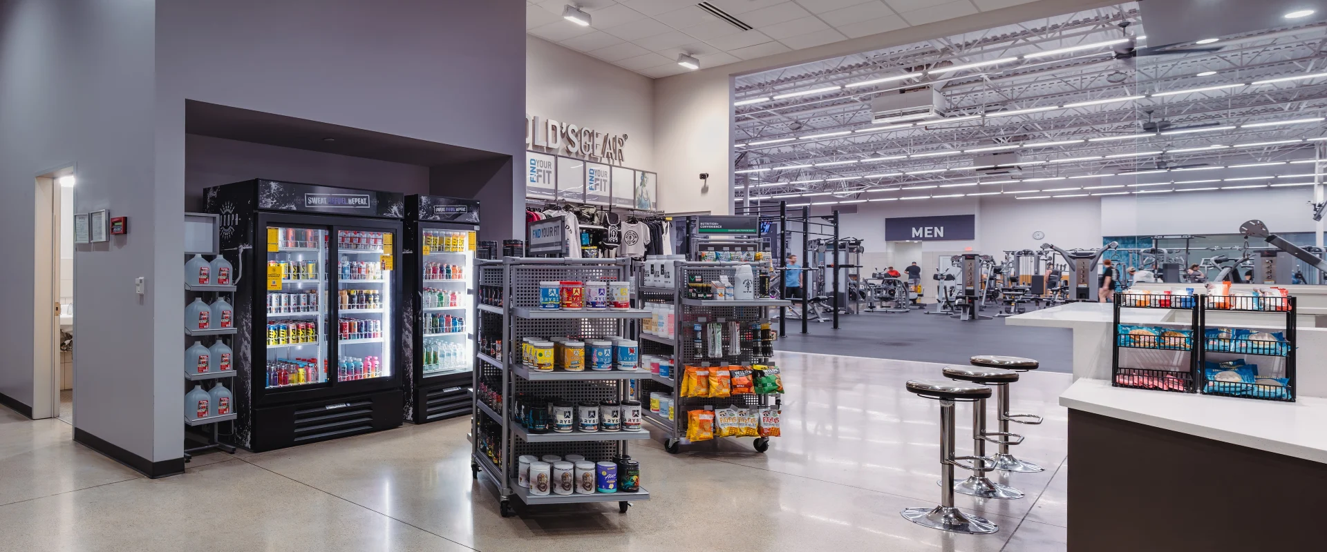 Pro Shop at Gold's Gym in Texas (Pflugerville), featuring refrigerated coolers stocked with energy drinks, protein shakes, and bottled water. Shelves display snacks, supplements, and branded merchandise. The sleek, modern area has polished floors, bar stools, and an open view of the gym floor.