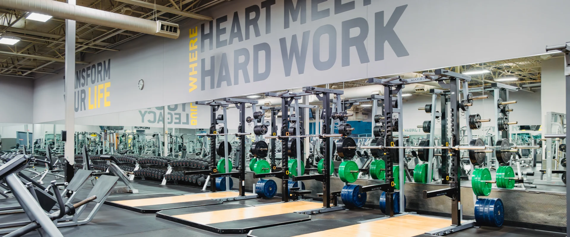 Olympic platforms at Gold's Gym Waco featuring weightlifting stations with Olympic bars, color-coded plates, and rubberized flooring. Mirrored walls and motivational text create a focused training environment, with bright overhead lighting illuminating the space.