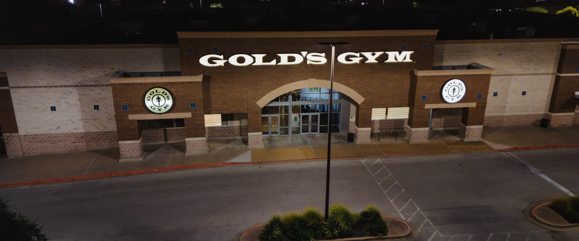 Nighttime view of Gold’s Gym Austin (Anderson Arbor) exterior featuring a large illuminated “Gold’s Gym” sign above the main glass entrance doors. The brick facade includes circular Gold’s Gym logos on the walls to the left and right of the entrance.
