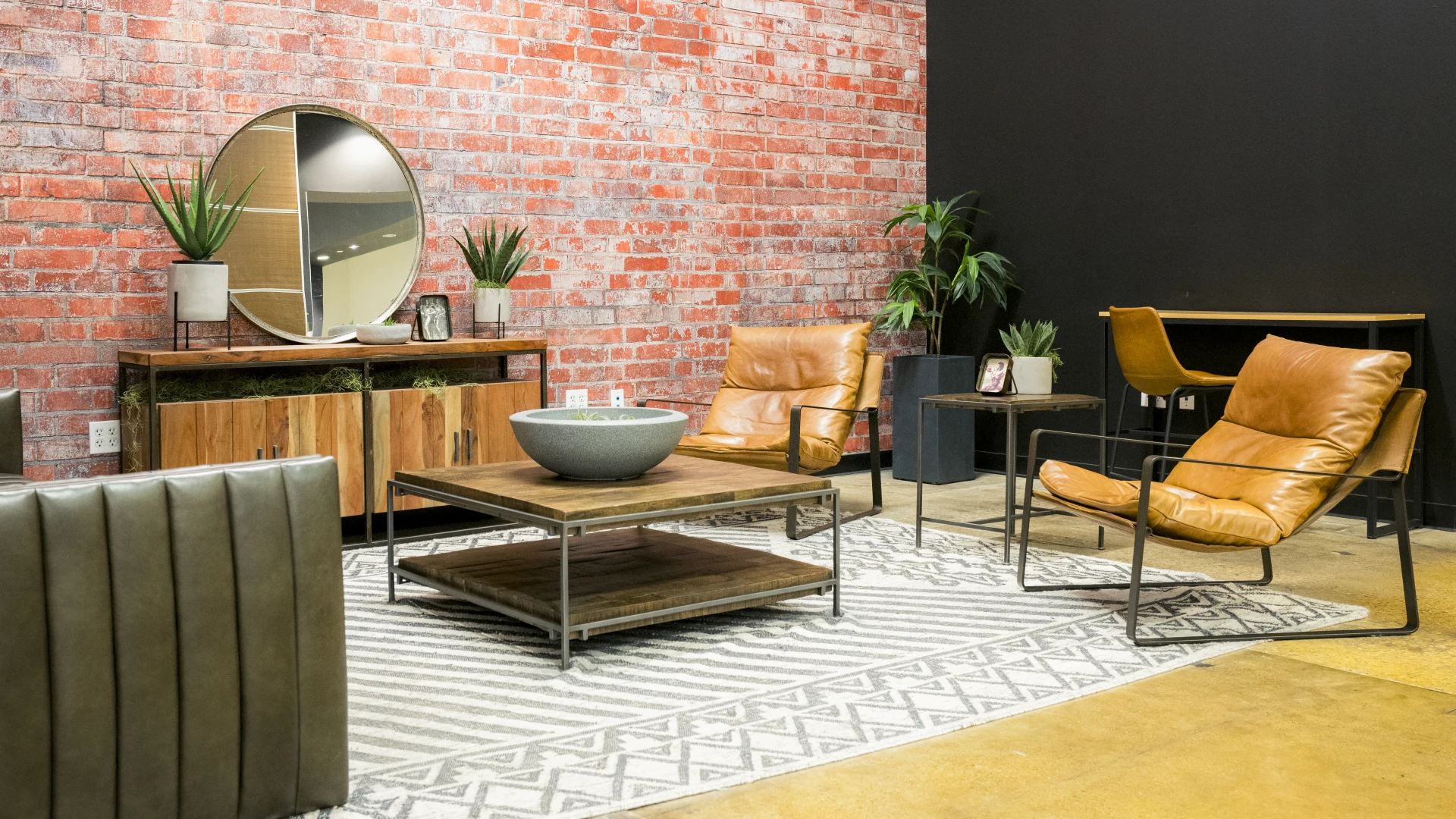 Lounge area at Gold's Gym Waxahachie featuring leather chairs, a wooden coffee table, and a matching credenza with a round mirror mounted on a red brick accent wall. The space includes potted plants, a patterned area rug, and a desk with additional seating, creating a stylish and comfortable environment for members.