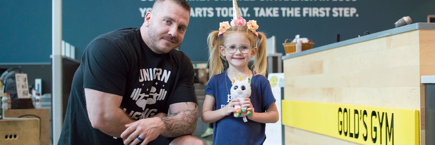 A man with tattoos and a young girl with a unicorn headband are standing together in what appears to be a classroom or educational setting, with a chalkboard and other school-related items visible in the background.