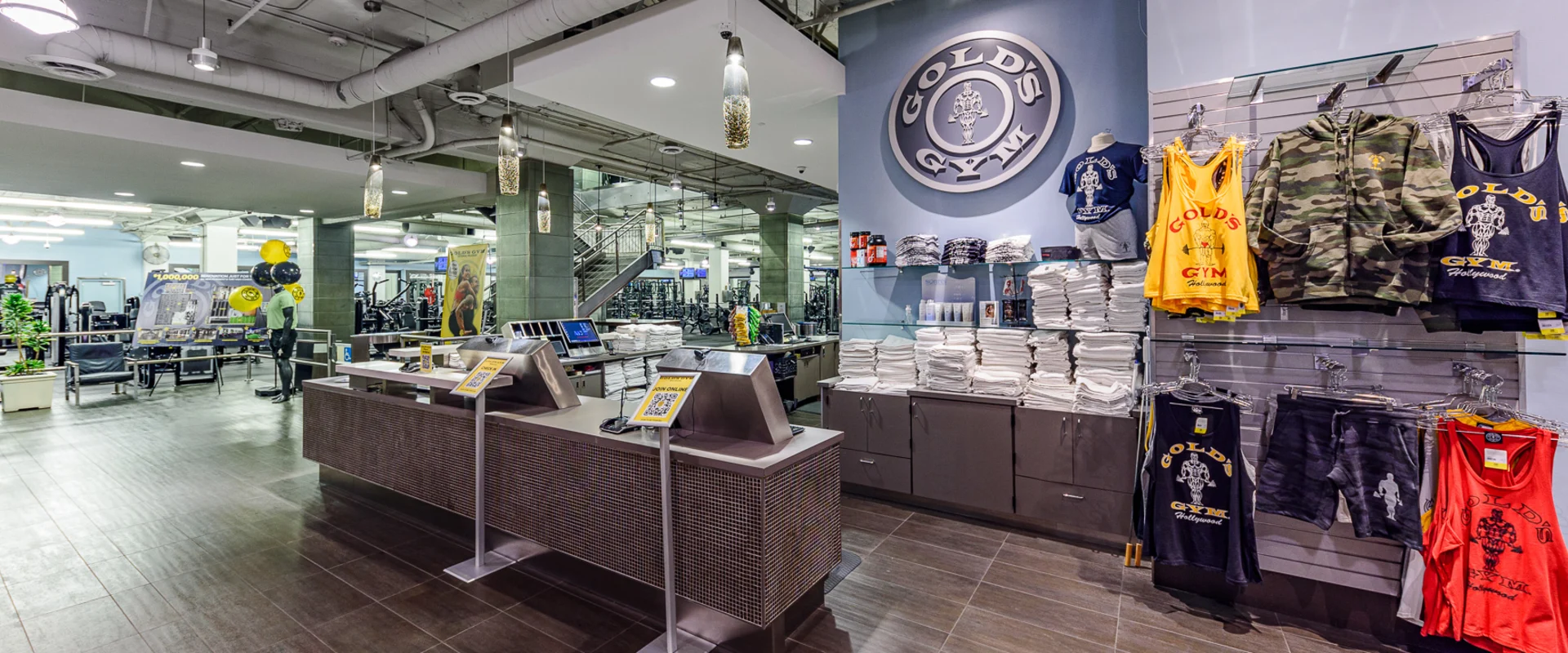 Front desk at Gold’s Gym Hollywood, featuring a modern reception counter with metal accents and touchscreen check-in stations. A large Gold’s Gym logo is mounted on a blue wall above shelves stocked with neatly folded white towels. To the right, gym apparel, including tank tops, hoodies, and shorts, is displayed on racks. Pendant lights hang from an exposed ceiling, illuminating the area. In the background, strength training equipment and a staircase leading to an upper level are visible.