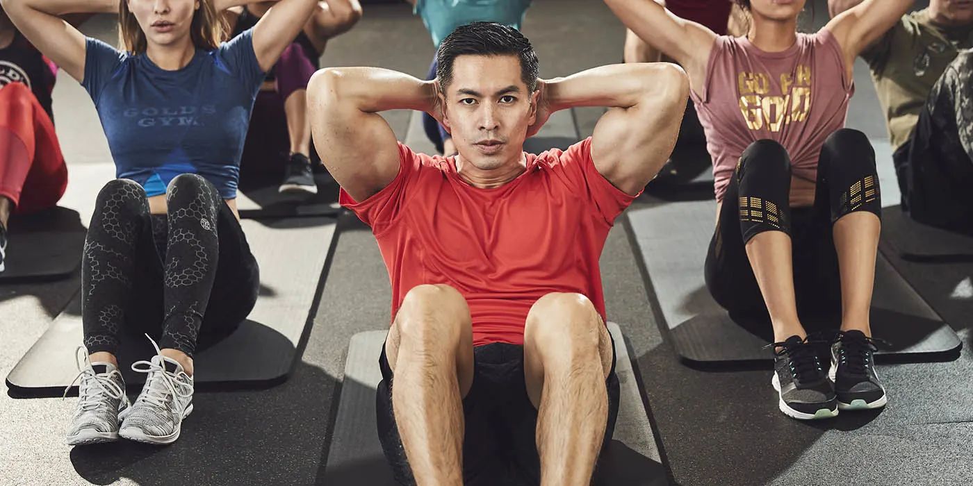 A group of people, including a man in a red shirt, are engaged in a fitness activity in a gym setting.