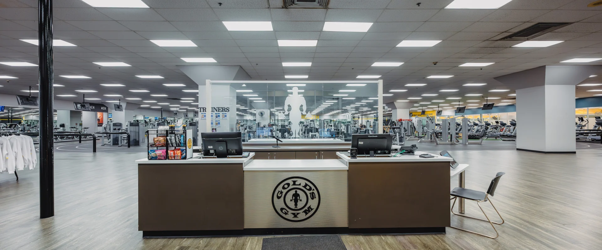 Front desk at Gold’s Gym in Austin (South Central), featuring a sleek reception counter with dual check-in stations and the Gold’s Gym logo displayed prominently on the front. Behind the desk, a large glass panel with the silhouette of a bodybuilder overlooks the gym floor. The area includes a display of snacks and supplements, with wood-style flooring and bright overhead lighting creating a welcoming entrance.