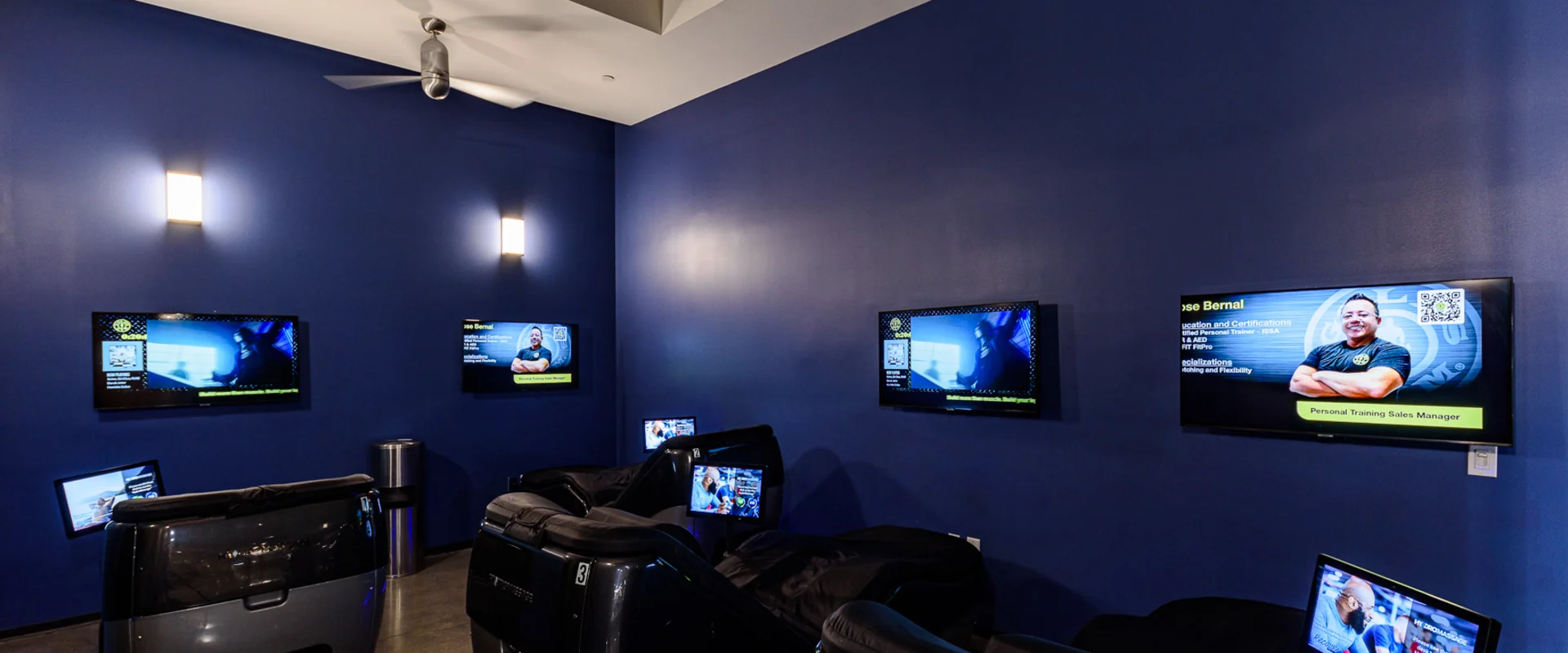 HydroMassage lounge area at Gold's Gym Garden Grove, featuring a row of black reclining massage chairs with individual screens for personalized relaxation settings. The dimly lit room has deep blue walls, modern wall sconces, and mounted TV screens displaying gym information and personal training promotions.