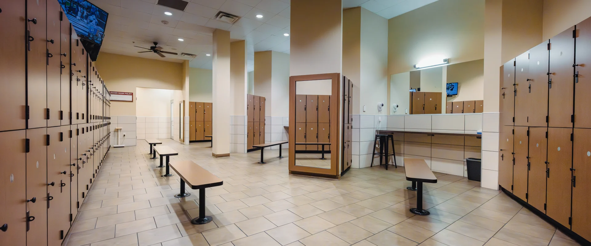 Locker room at Gold’s Gym Austin (Anderson Arbor) with beige tiled flooring, wooden lockers lining the walls, and benches in the center. A full-length mirror, a grooming area with sinks, mirrors, and hair dryers, and a wall-mounted TV are visible. Ceiling fans and recessed lighting illuminate the space.