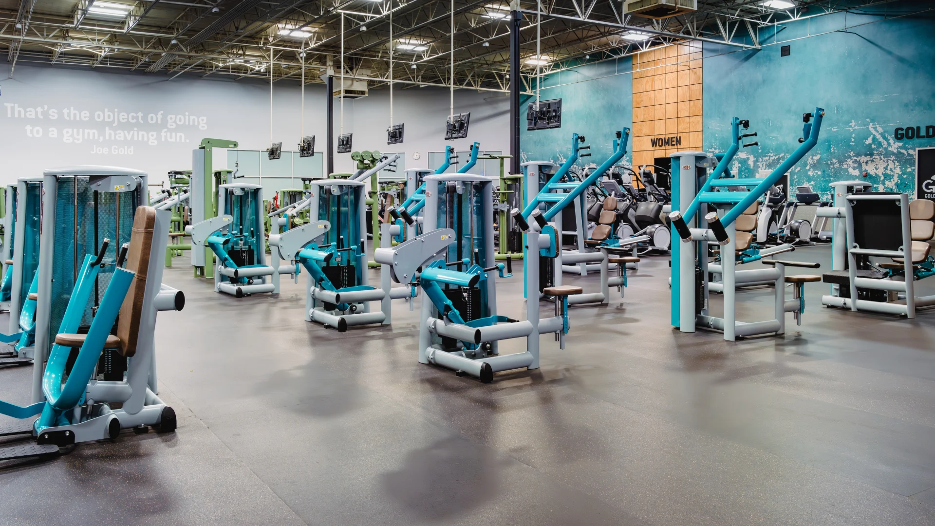 Wide gym floor with multiple strength machines under high ceilings and bright lighting.