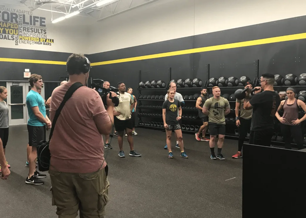 A group of people, both men and women, are gathered in a fitness center or gym, with various exercise equipment and motivational signage visible in the background.