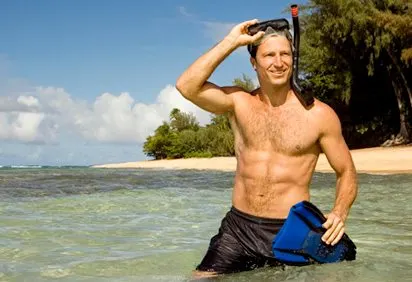 A shirtless man stands in shallow water, holding a snorkel mask and gazing towards the camera, with a tropical beach and lush vegetation visible in the background.