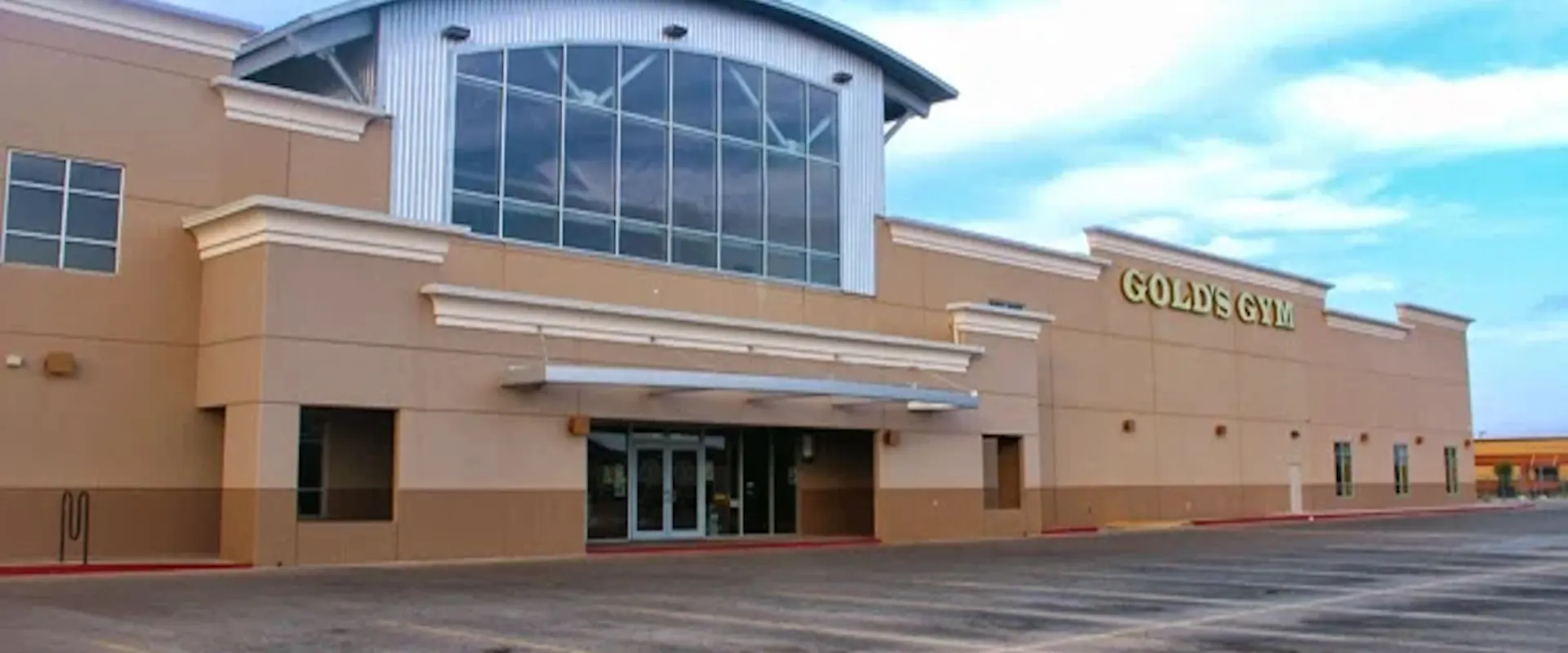 Exterior of Gold’s Gym McAllen with tan facade, large windows, and yellow signage on a sunny day.
