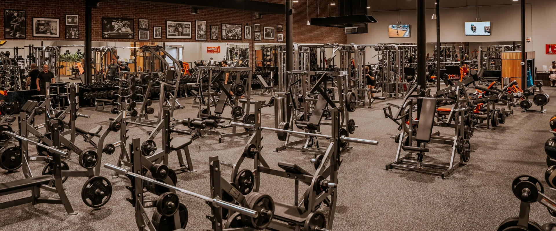 Gold's Gym Memphis, Tennessee (White Station) free weight area featuring multiple squat racks, bench presses, and plate-loaded machines arranged across a spacious floor. Dumbbells are neatly racked along the back wall beneath framed black-and-white bodybuilding photos on an exposed brick wall. Several gym-goers are seen lifting weights and using strength equipment. Overhead lighting highlights the industrial-style ceiling with black beams and exposed ductwork. Flat-screen TVs are mounted on the walls, providing entertainment.