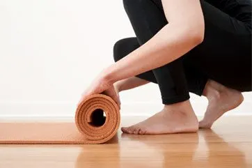 A person's legs and feet are visible, with a rolled-up orange yoga mat on the floor in the foreground, and a plain white background behind them.